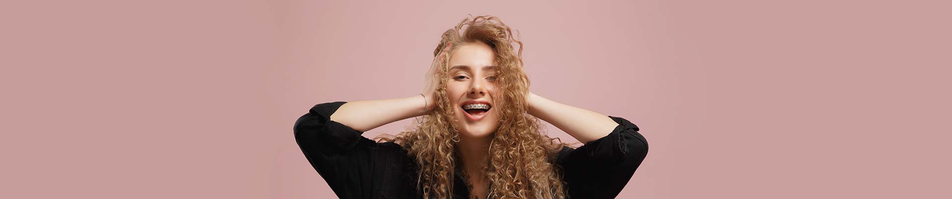 Cheerful beautiful girl with curly hair, braces on her teeth, holds her hands on her head, smiling broadly with her mouth open.