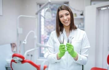 Smiling female dentist in white coat holding dental instruments in green gloves.