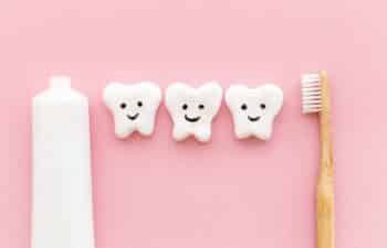 Three smiling tooth characters with toothpaste and bamboo toothbrush on pink background.