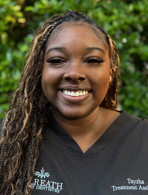 Smiling woman with braided hair wearing Gilreath Dentistry uniform against green foliage background.