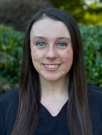 Woman smiling at camera outdoors with green foliage background, professional headshot.