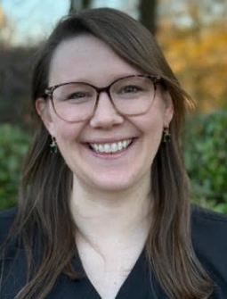 Young woman with glasses smiling at camera outdoors with blurred autumn foliage background.