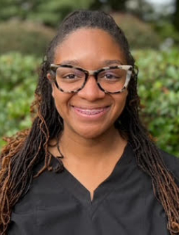 Portrait of smiling woman with glasses wearing black scrubs against green foliage background.